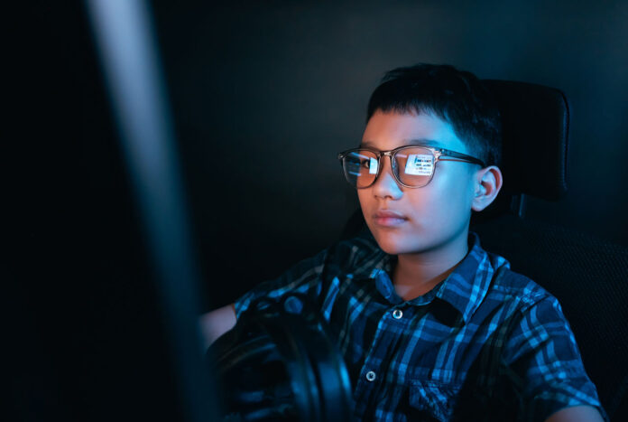 Young kid on the computer playing games, watching entertainment or browsing the online digital internet, wearing glasses and dark background theme.